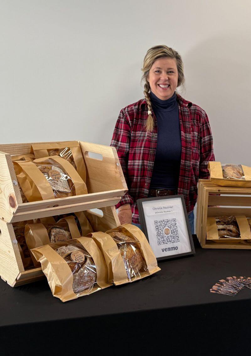 A smiling woman in a red plaid shirt stands behind a table displaying loaves of bread packaged in brown paper bags with clear windows. The bread is arranged in wooden crates, and a framed Venmo QR code for payment is placed on the table