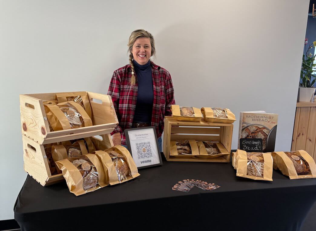 A smiling woman in a red plaid shirt stands behind a table displaying loaves of bread packaged in brown paper bags with clear windows. The bread is arranged in wooden crates, and a framed Venmo QR code for payment is placed on the table
